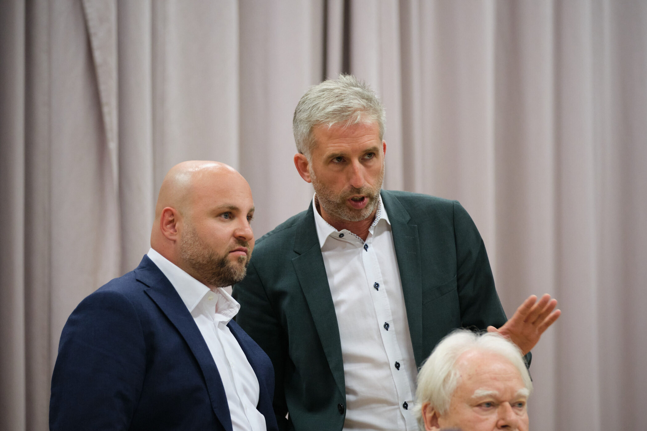 Drei Männer in Anzügen, einer spricht, vor einem Vorhang.