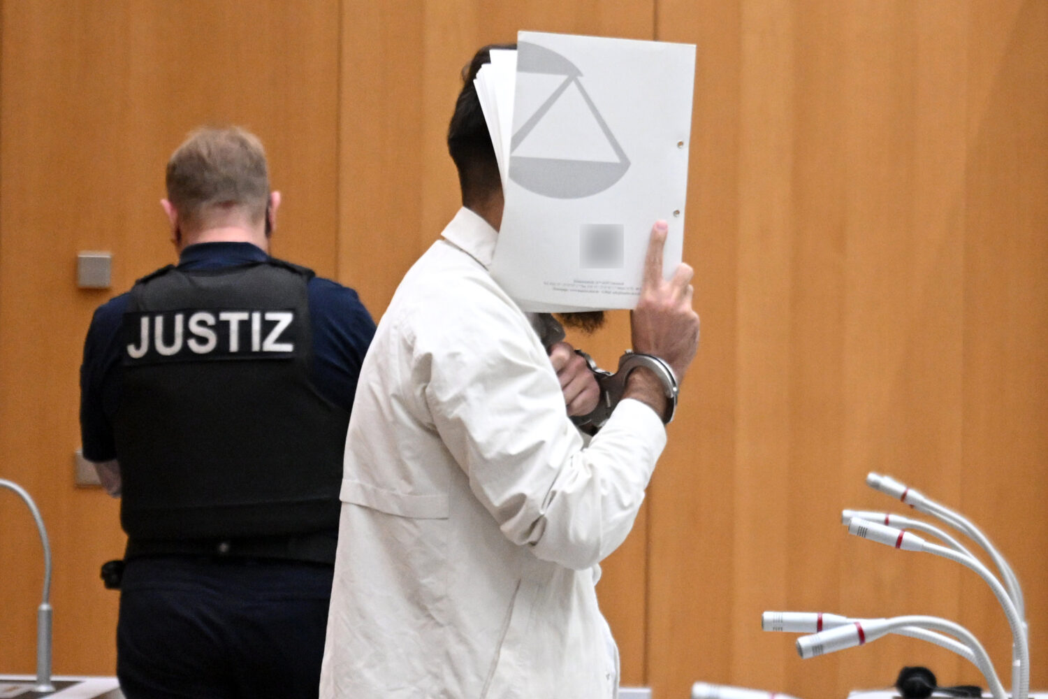 Zwei Personen im Raum, eine mit Handschellen, hält ein Dokument vor das Gesicht.