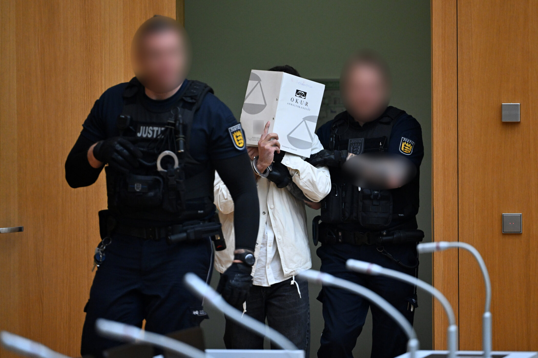 Drei Personen, eine hält ein Dokument vor das Gesicht, zwei in Uniform.