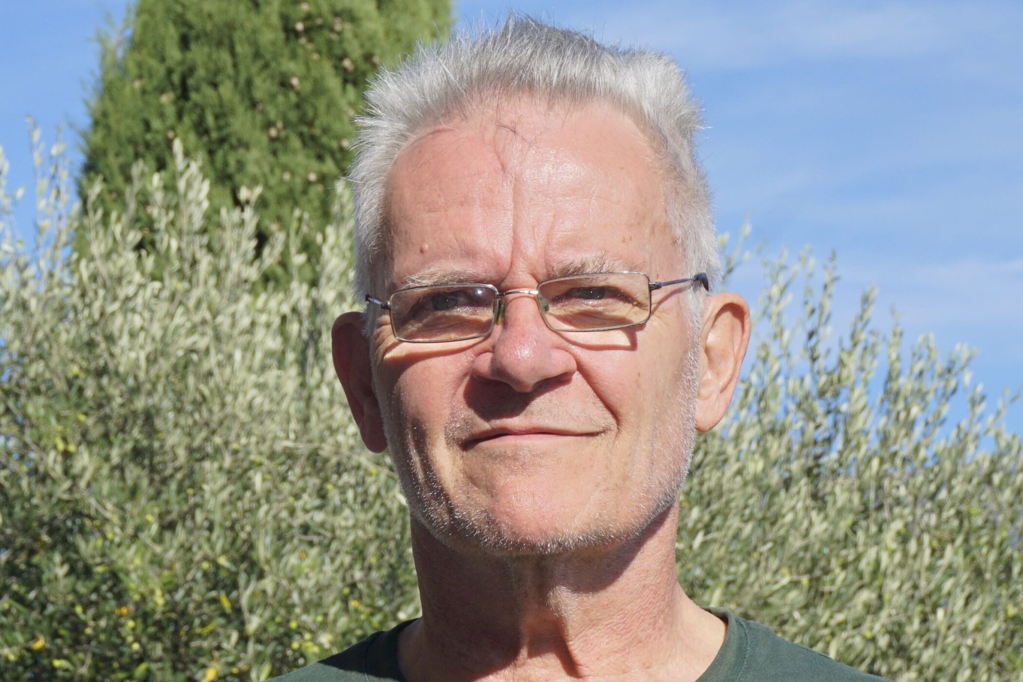 Älterer Mann mit Brille vor Bäumen und blauem Himmel.