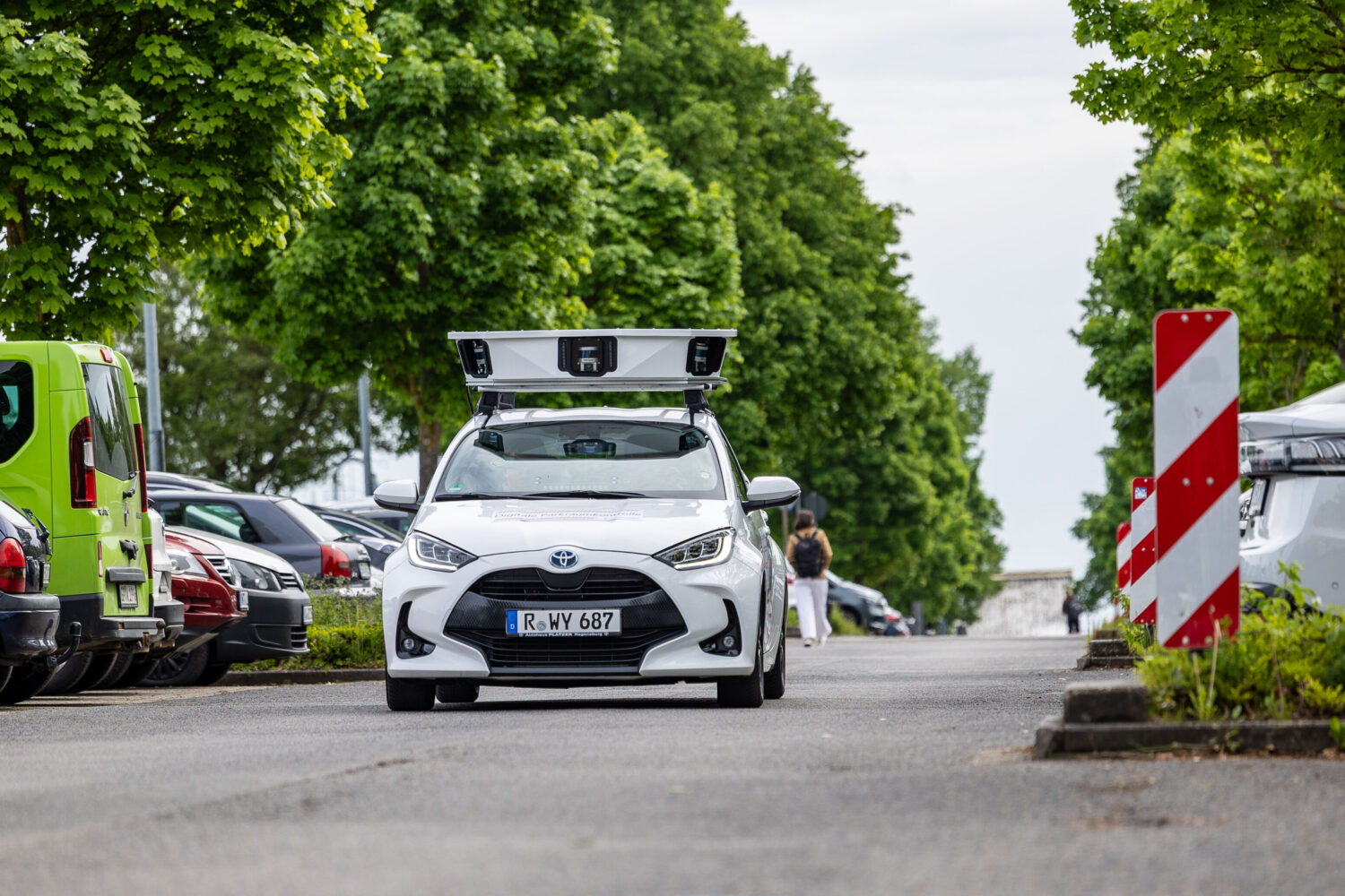 Weißes Auto mit Dachaufbau auf Straße zwischen geparkten Autos und Bäumen.