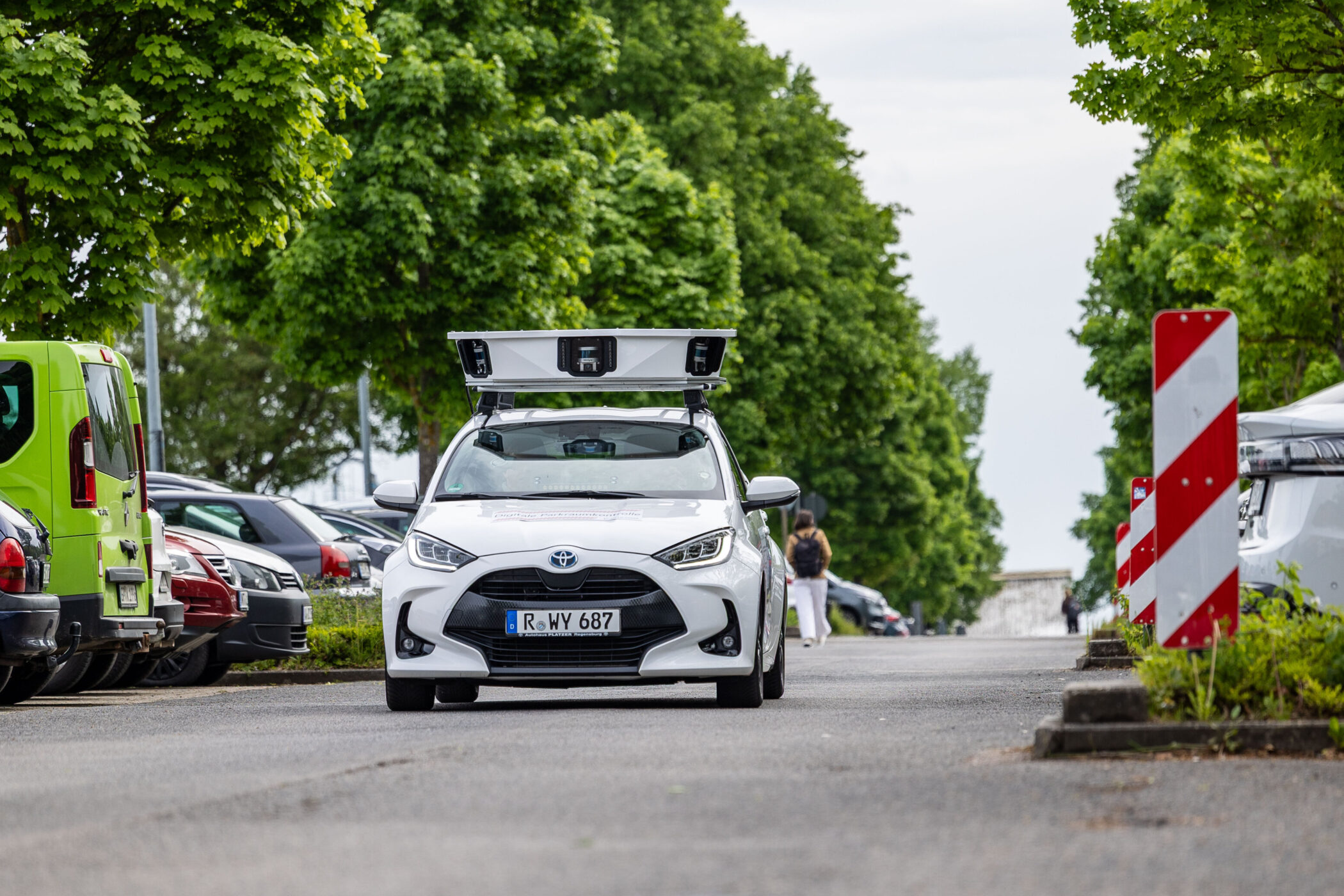 Weißes Auto mit Dachaufbau auf Straße zwischen geparkten Autos und Bäumen.