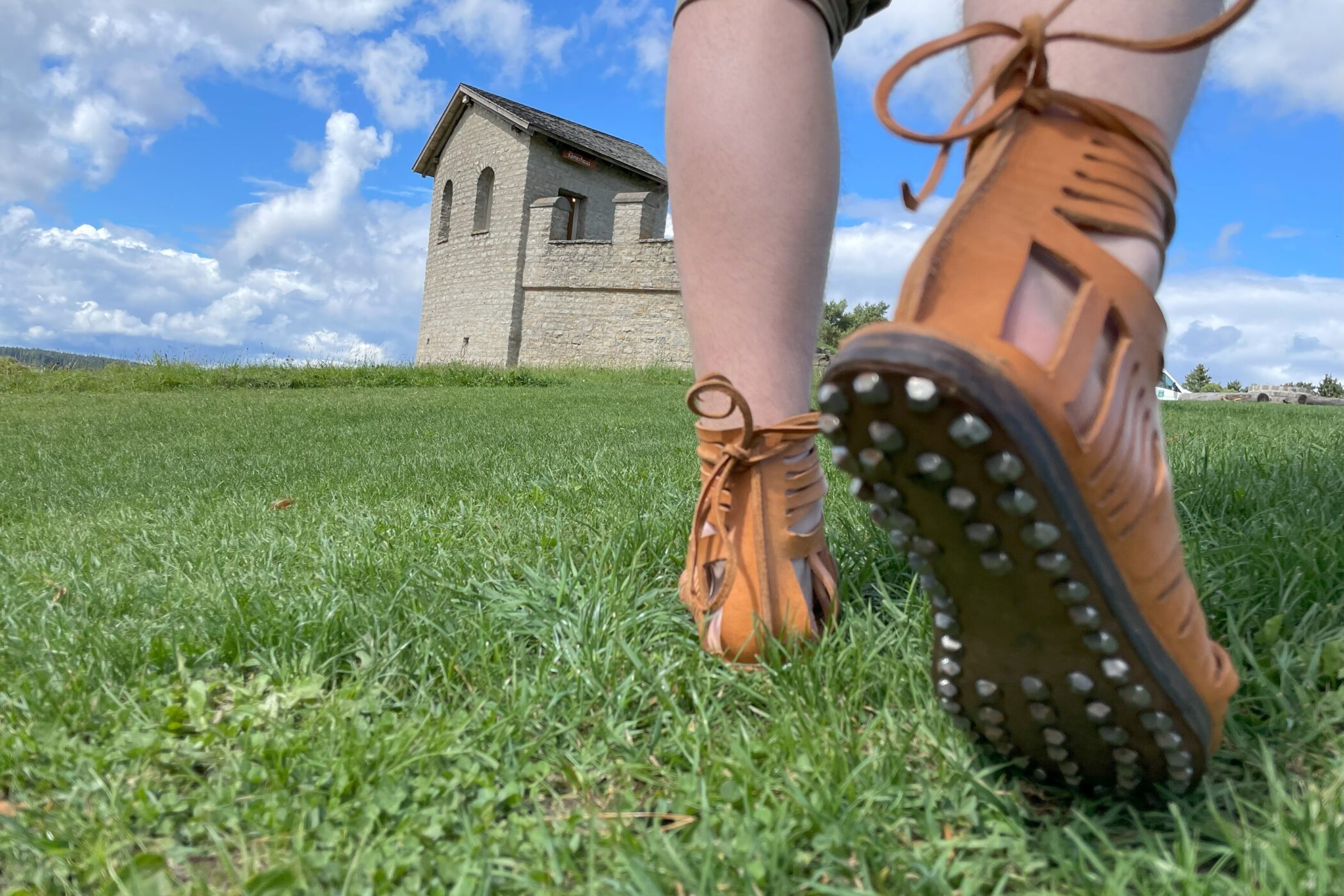 Beine in Sandalen auf Gras, im Hintergrund ein kleines Steingebäude.