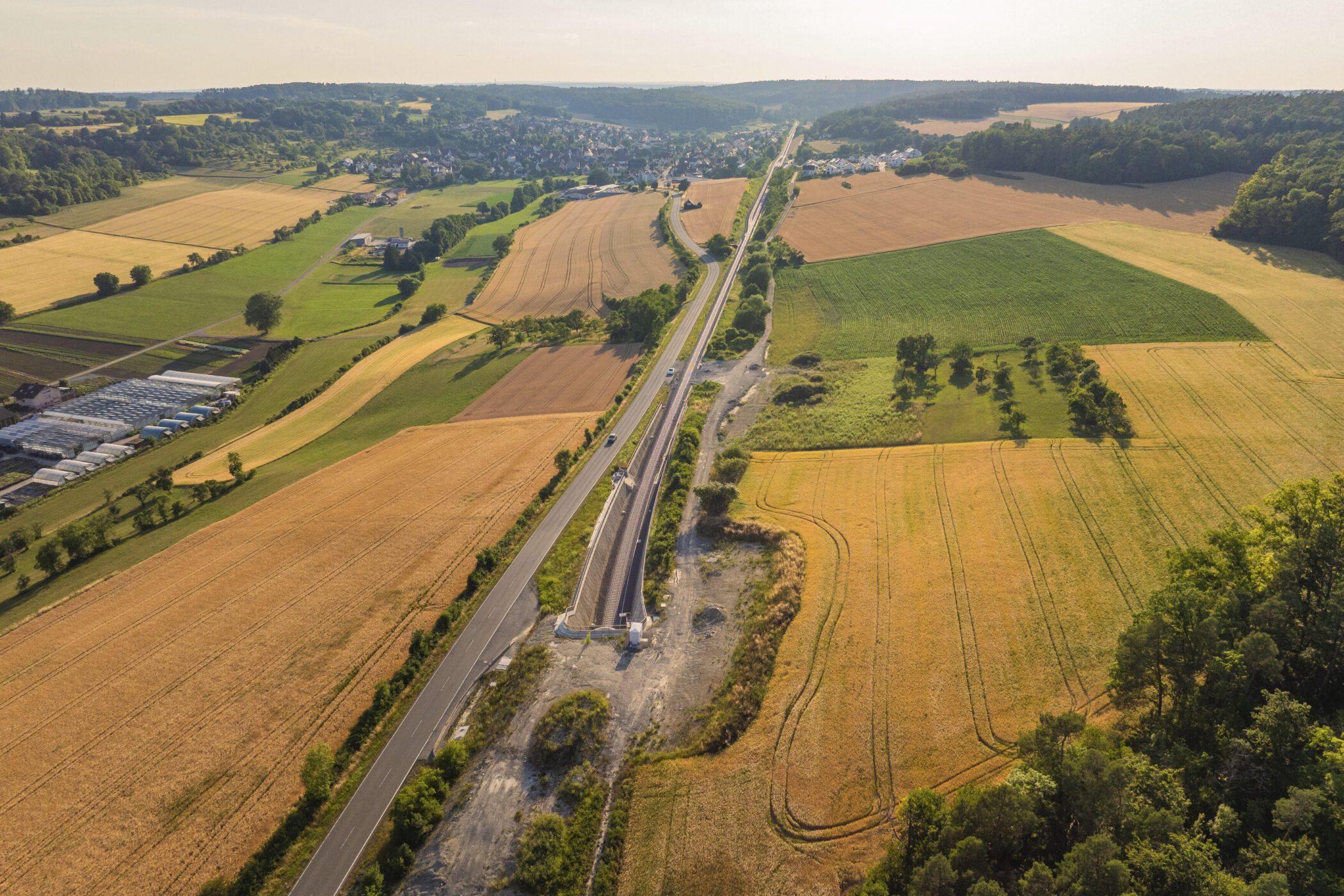 Luftaufnahme von Feldern, Straße und Schienen, im Hintergrund ein Dorf.