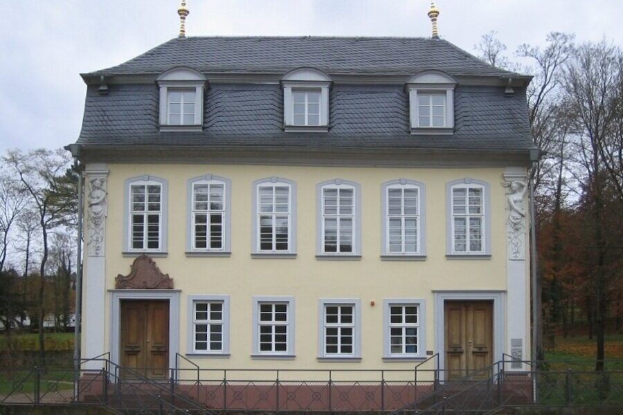 Gelbes zweistöckiges Gebäude mit grauem Dach und mehreren Fenstern, umgeben von Bäumen.