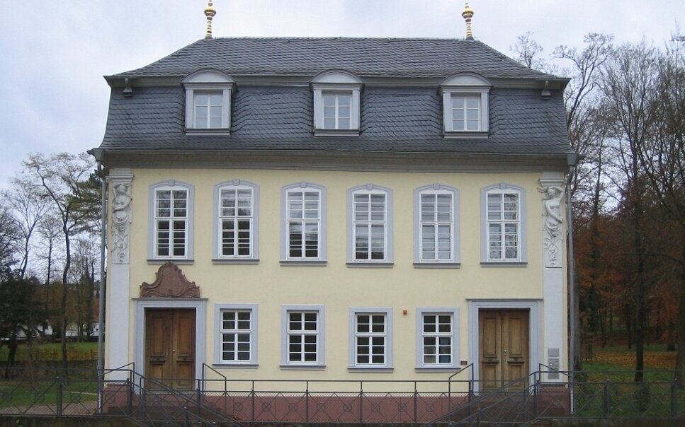 Gelbes zweistöckiges Gebäude mit grauem Dach und mehreren Fenstern, umgeben von Bäumen.