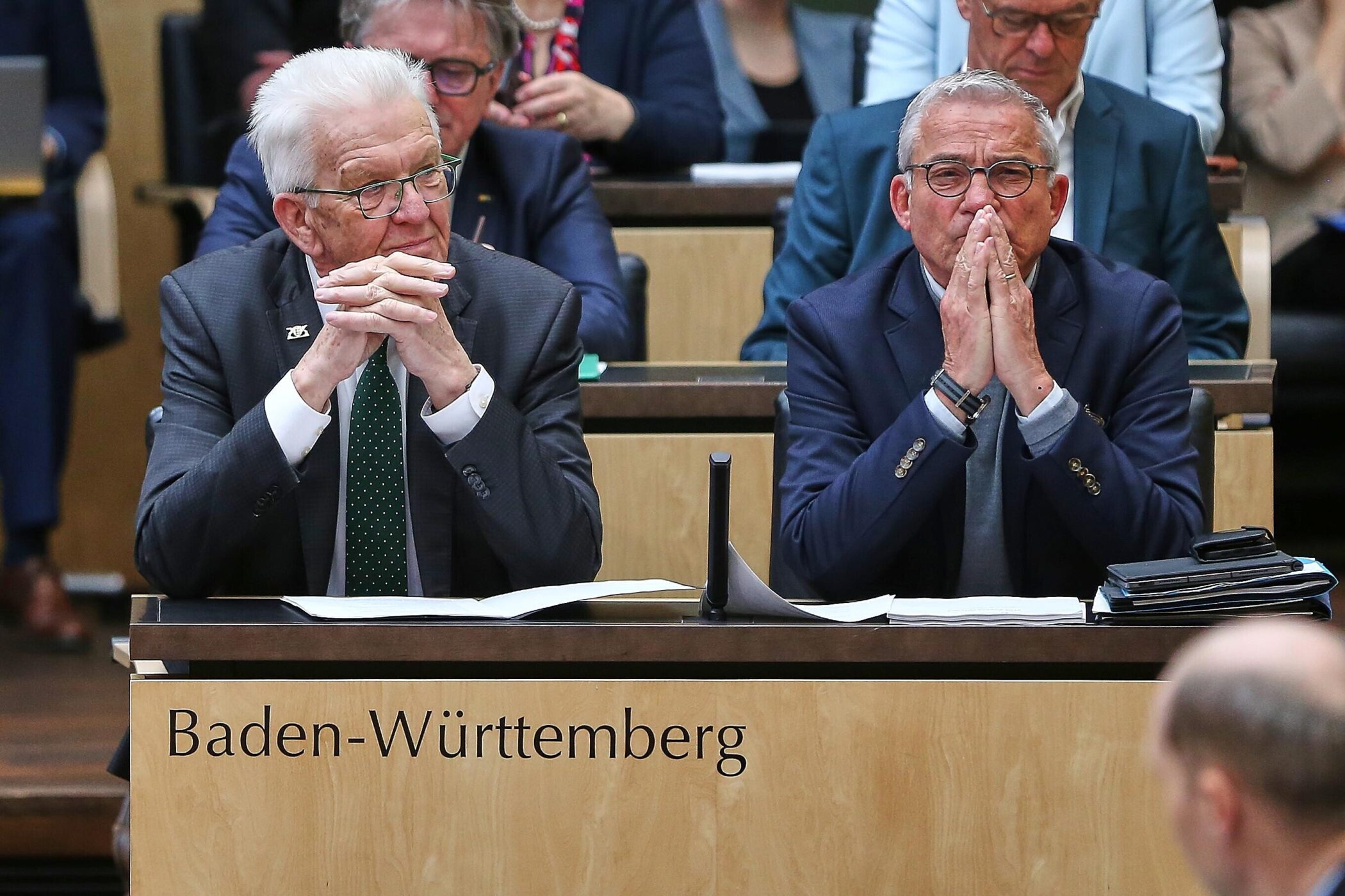 Zwei Männer sitzen in einem Saal, Schild mit "Baden-Württemberg".