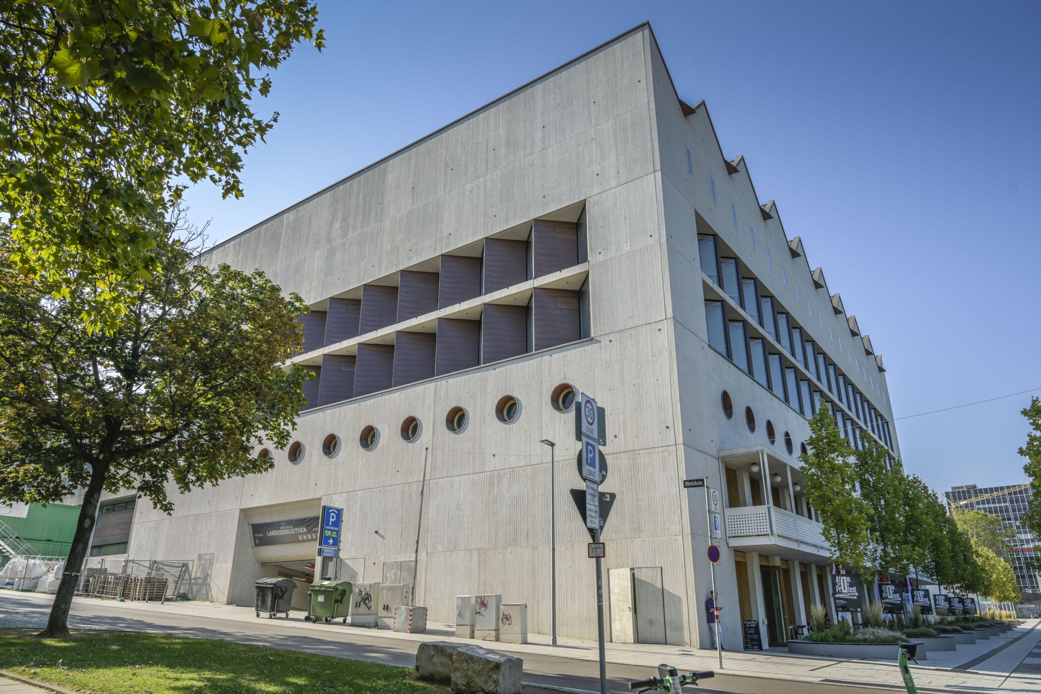 Modernes Gebäude mit runden Fenstern, Bäumen und Verkehrsschildern im Vordergrund.
