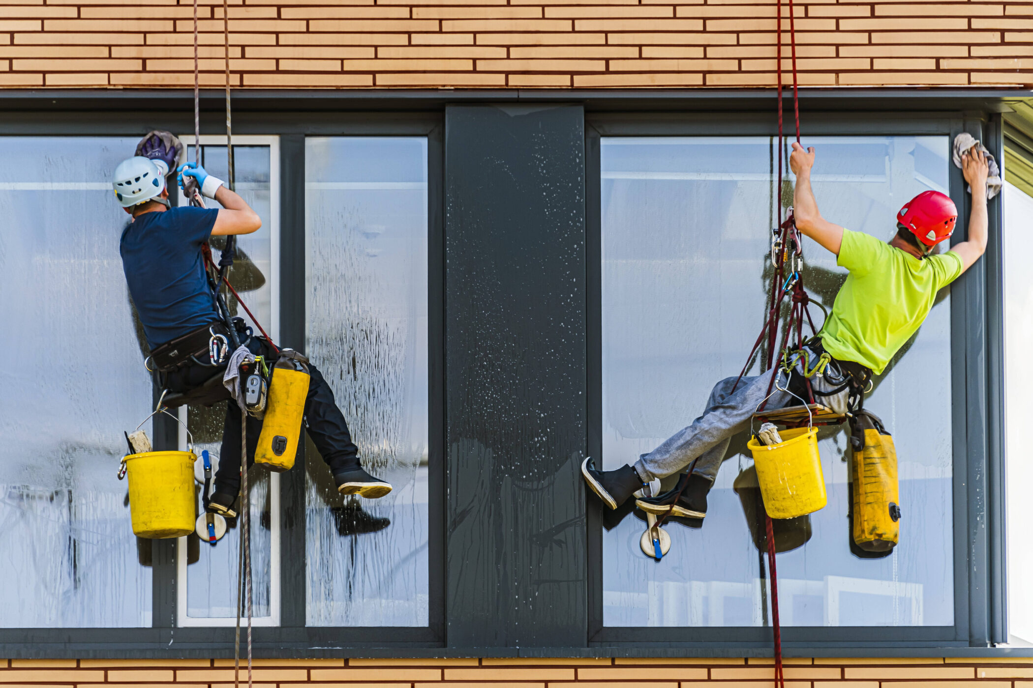Zwei Personen reinigen Fenster eines Gebäudes, gesichert mit Seilen und Helmen.