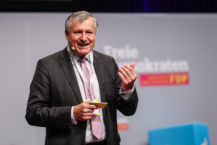 Älterer Mann im Anzug spricht auf einer Bühne, Mikrofon am Ohr.