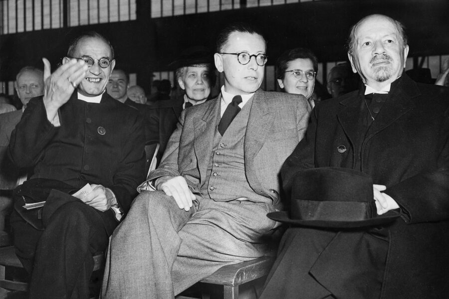 Drei Männer in Anzügen sitzen nebeneinander, umgeben von weiteren Personen.