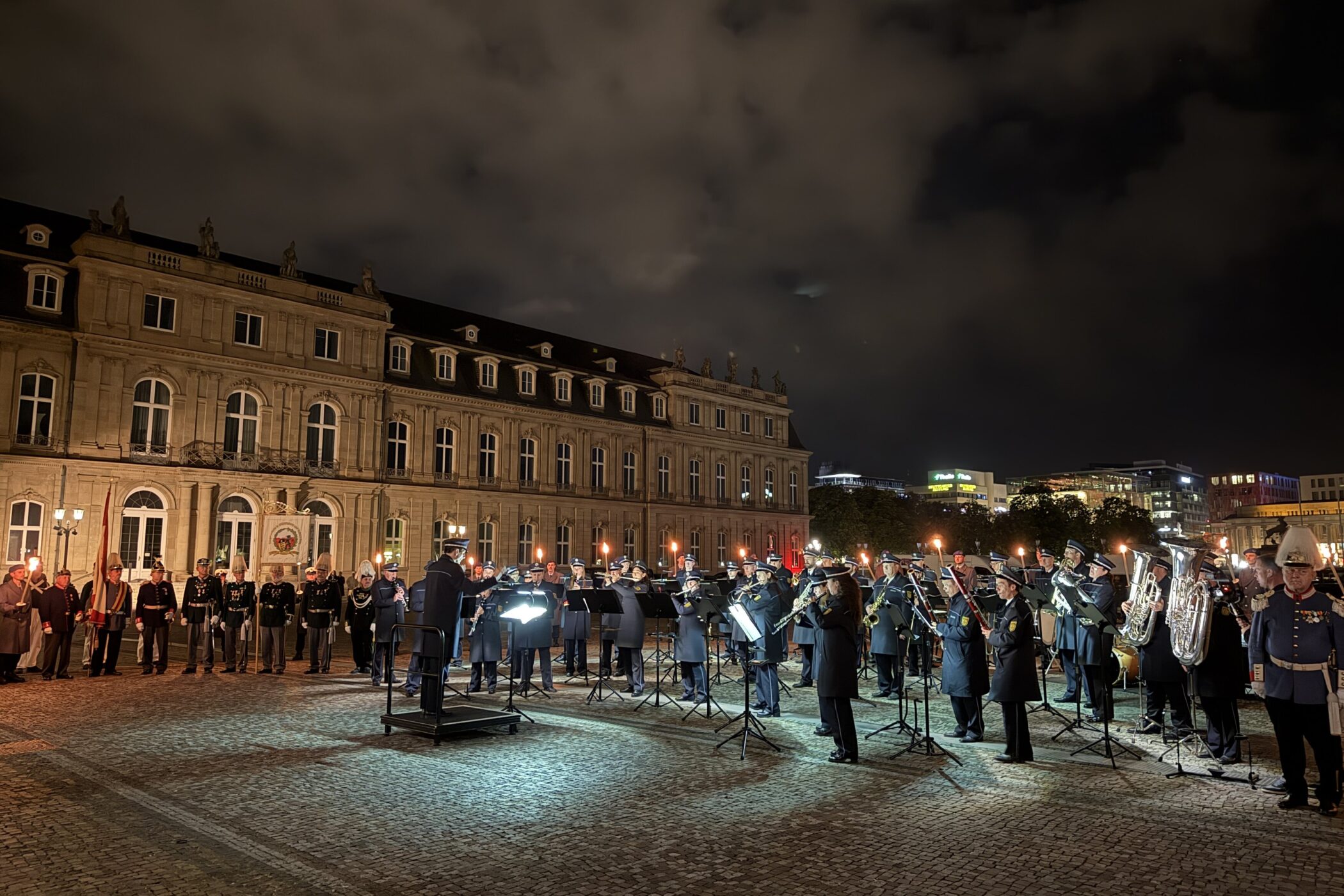 Menschen in Uniformen mit Fackeln und Musikinstrumenten vor einem beleuchteten Gebäude.