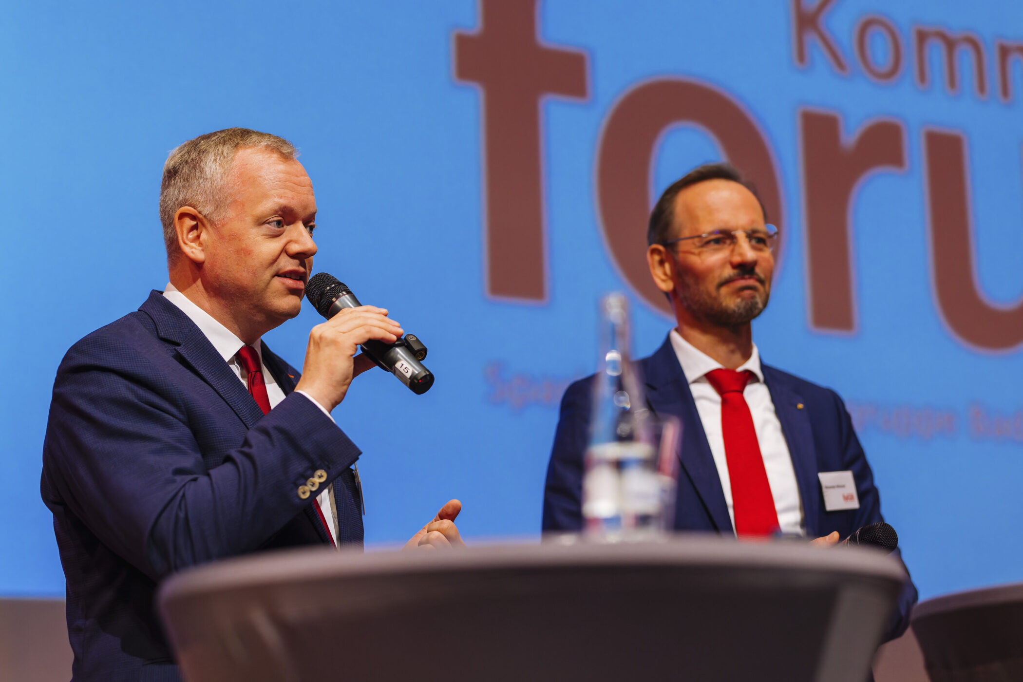 Zwei Männer in Anzügen auf einer Bühne, einer spricht in ein Mikrofon.