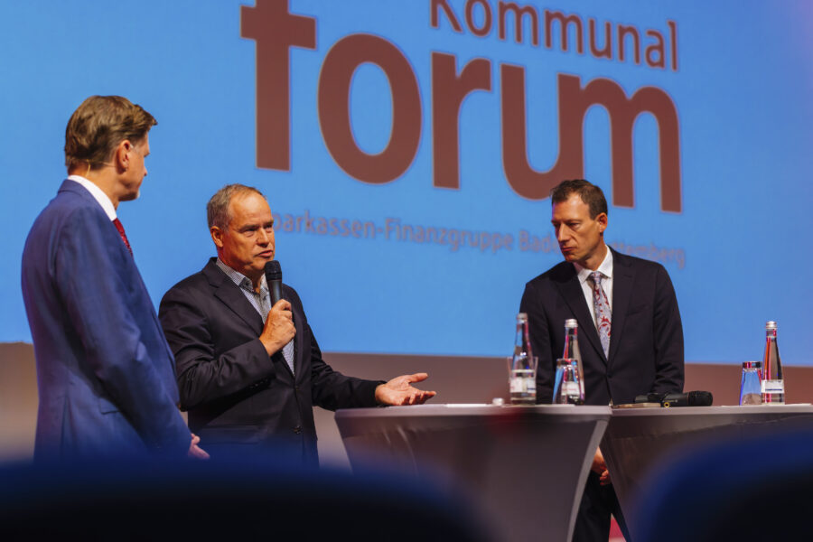 Drei Männer in Anzügen auf einer Bühne, einer spricht in ein Mikrofon.