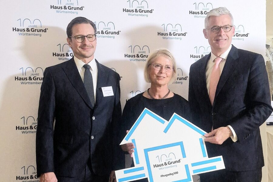 Drei Personen in Anzügen vor "Haus & Grund Württemberg"-Hintergrund. Frau hält Schild.