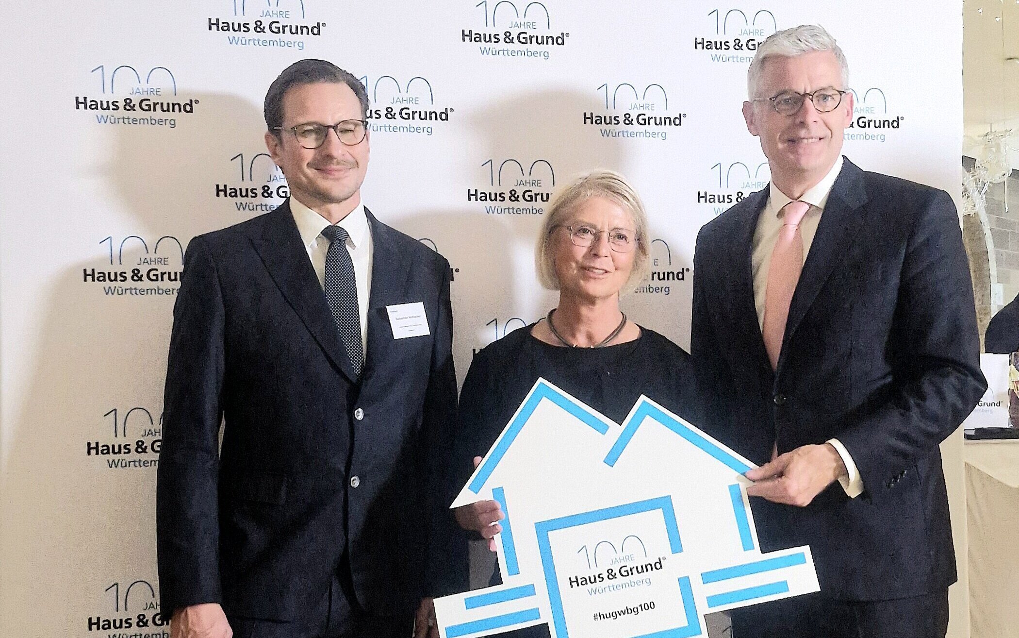 Drei Personen in Anzügen vor "Haus & Grund Württemberg"-Hintergrund. Frau hält Schild.