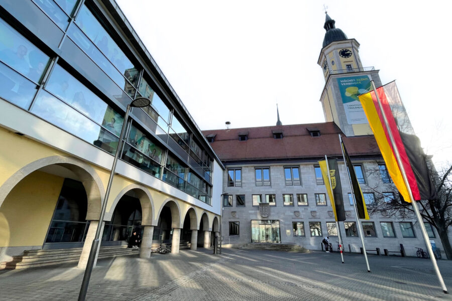 Gebäude mit Arkaden, Turm mit Uhr, mehrere Flaggen, gepflasterter Platz.