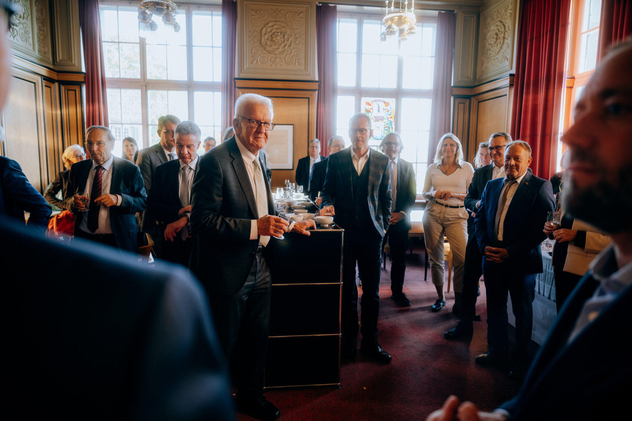 Mehrere Personen in Anzügen stehen in einem Raum mit großen Fenstern.
