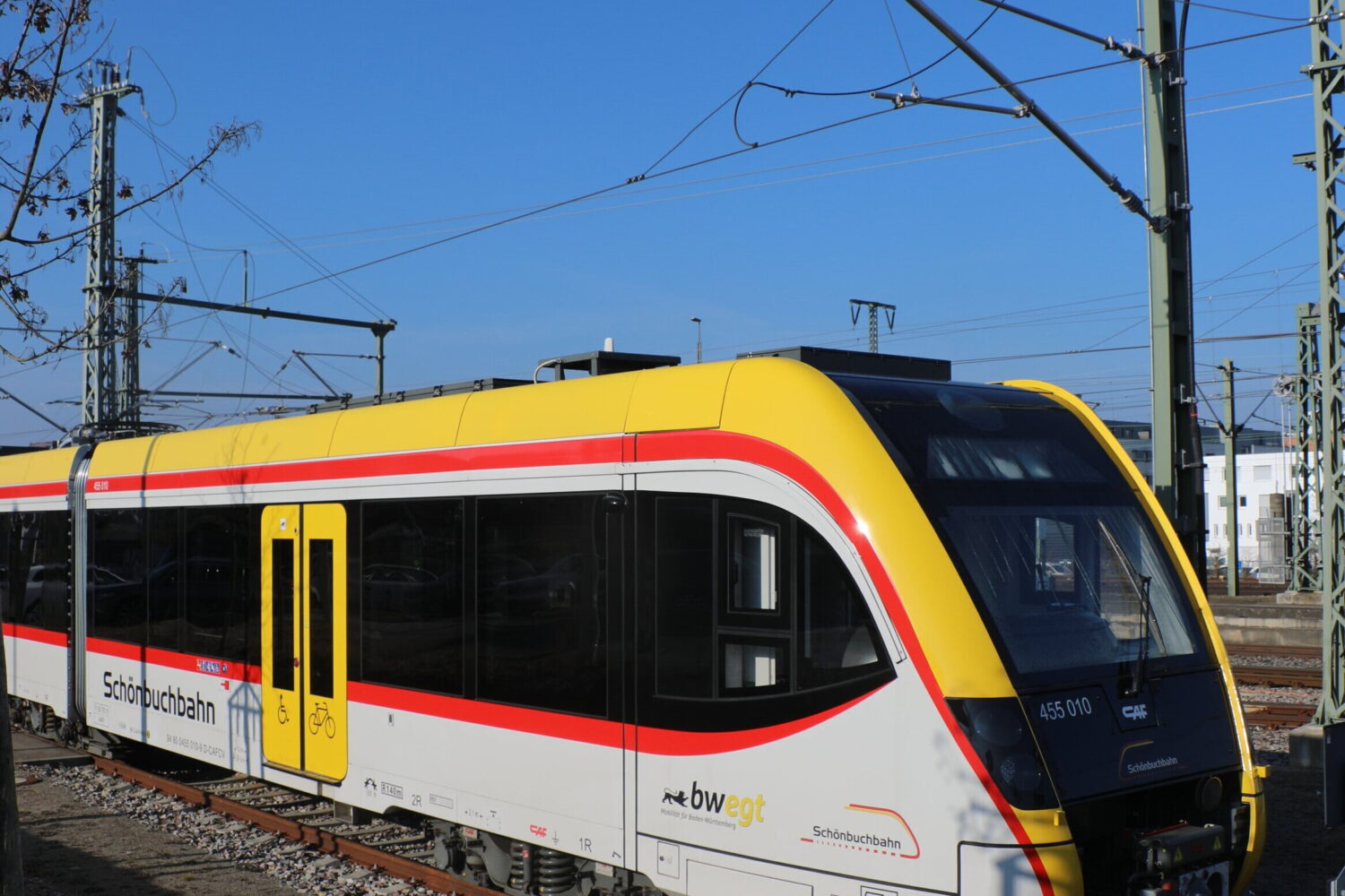 Gelb-weißer Zug mit rotem Streifen auf Gleisen, blauer Himmel im Hintergrund.