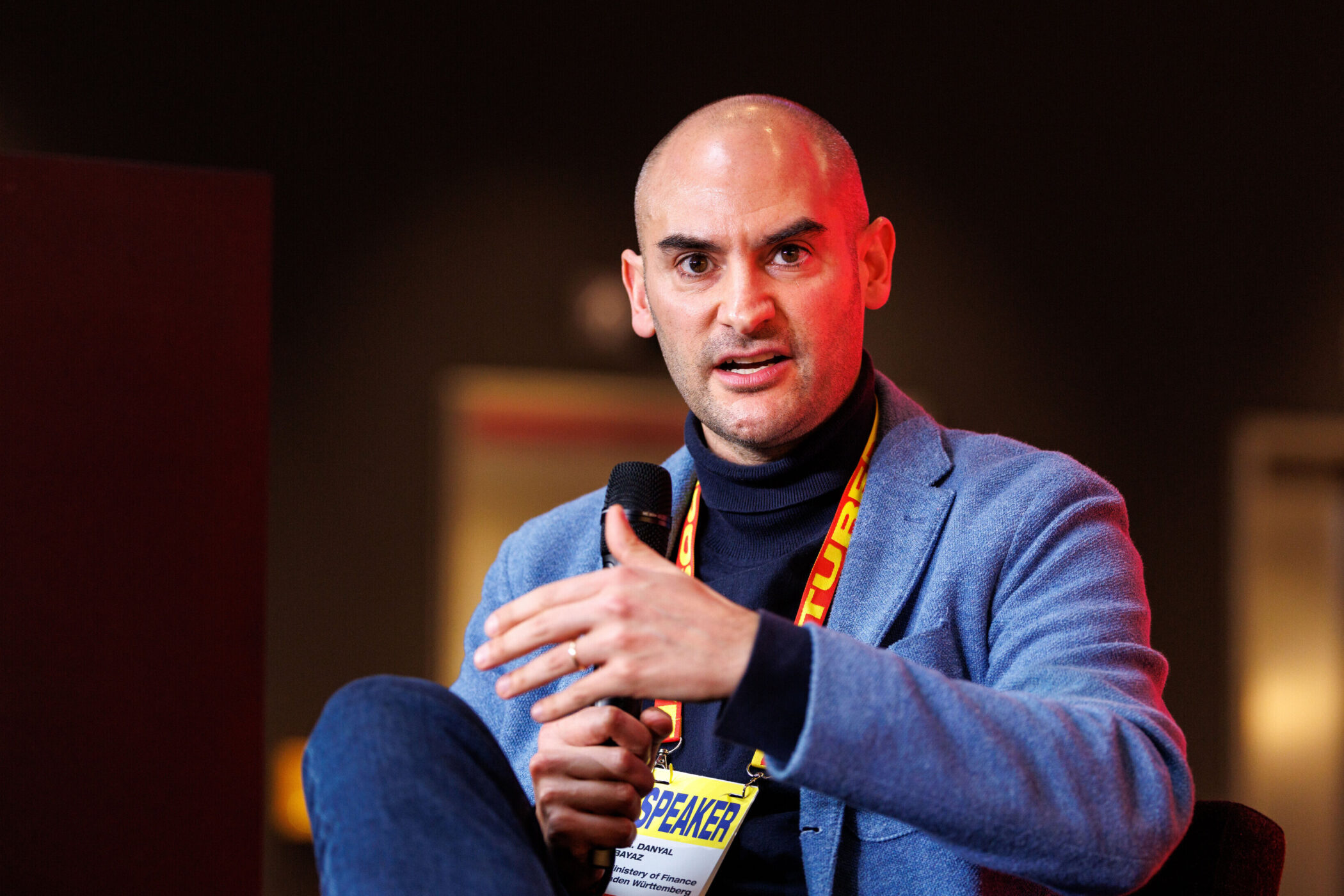 Mann mit Mikrofon und Namensschild spricht, trägt blauen Blazer.