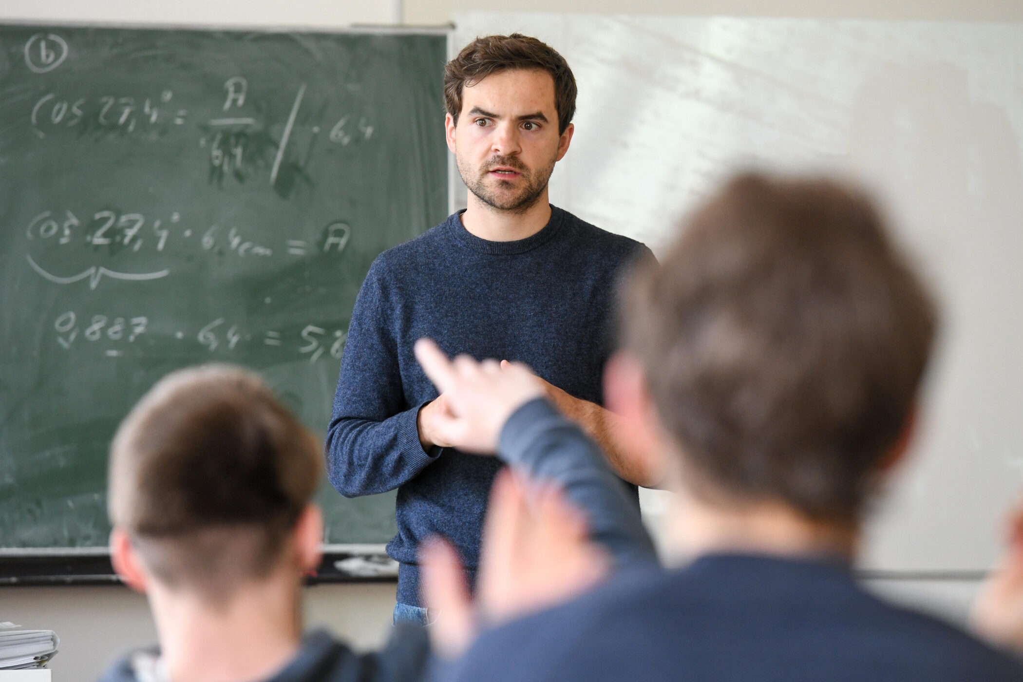 Ein Mann steht vor einer Tafel, Schüler heben die Hand.