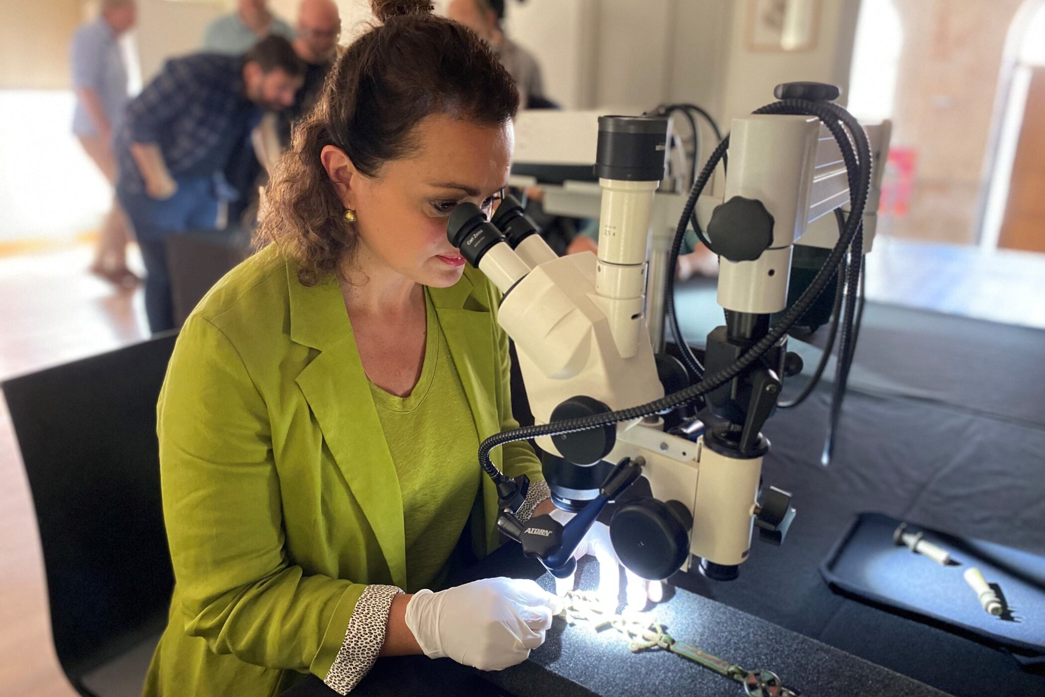 Frau untersucht Objekt unter Mikroskop, trägt grünen Blazer und Handschuhe.
