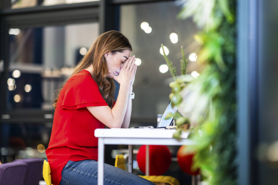 Frau in rotem Oberteil sitzt am Tisch vor Laptop, hält Hände vor Gesicht.