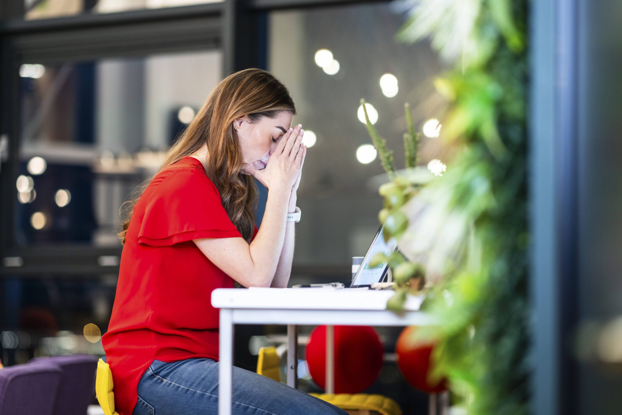 Frau in rotem Oberteil sitzt am Tisch, Hände vor dem Gesicht, Laptop vor ihr.