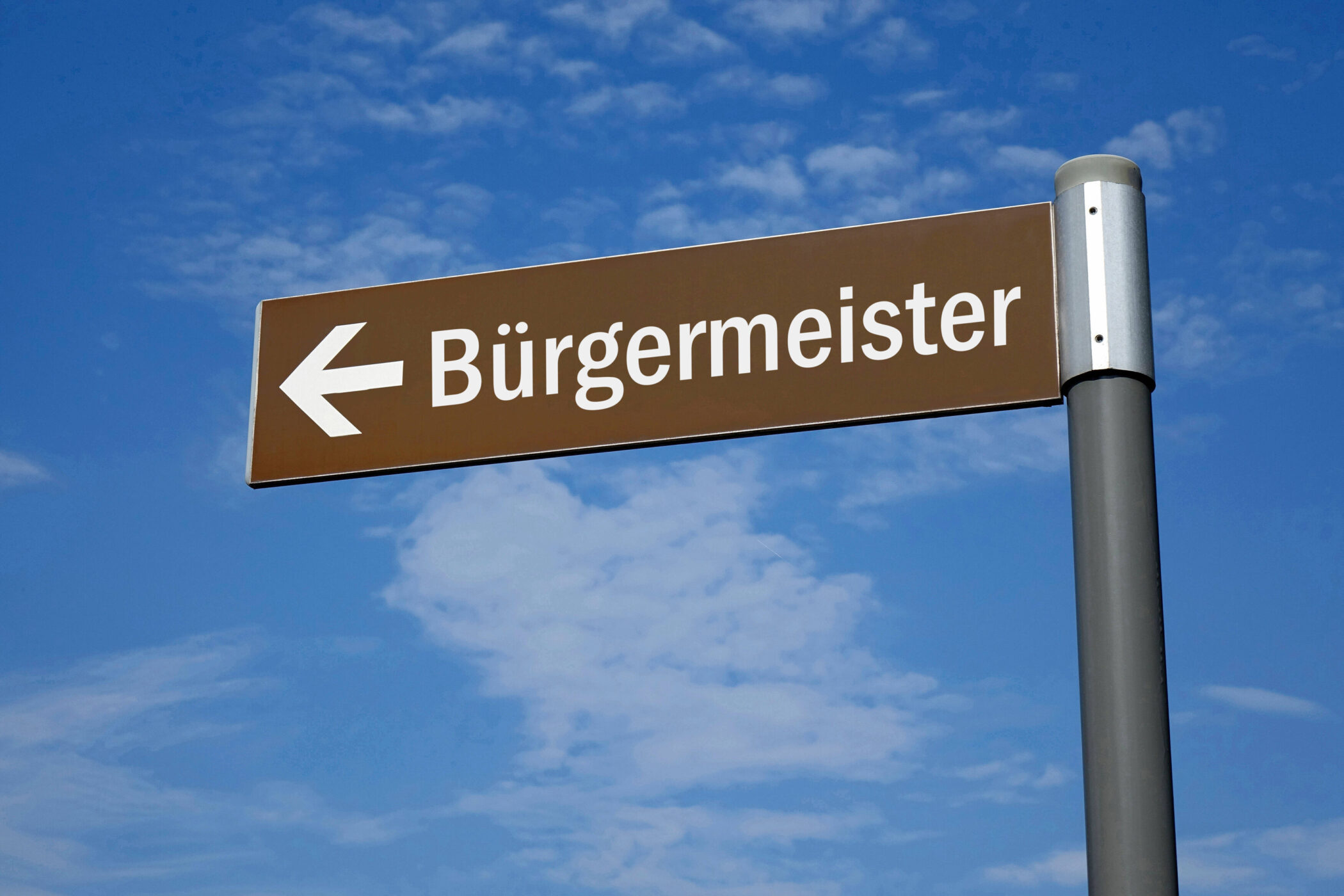 Braunes Schild mit Pfeil und Text "Bürgermeister" vor blauem Himmel.