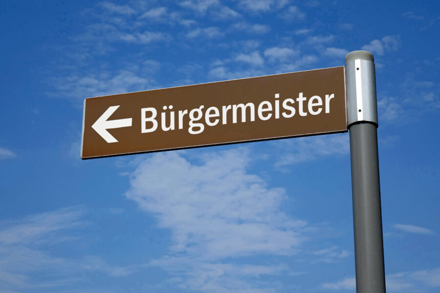 Braunes Schild mit Pfeil und Text "Bürgermeister" vor blauem Himmel.