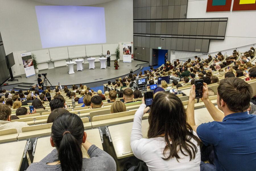 Großer Hörsaal mit vielen Menschen, einige filmen mit Smartphones, Vortragende vorne.
