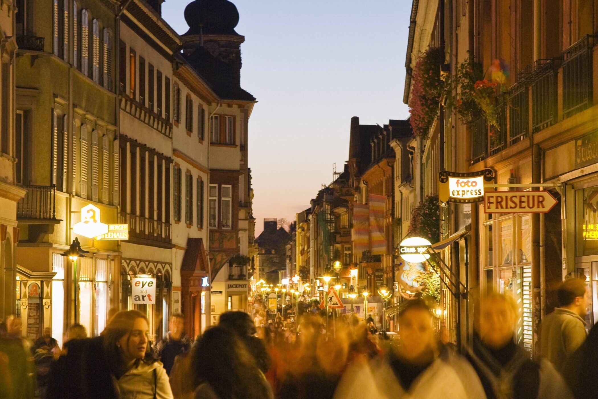 Belebte Straße bei Dämmerung, viele Menschen, Geschäfte mit Leuchtreklamen.