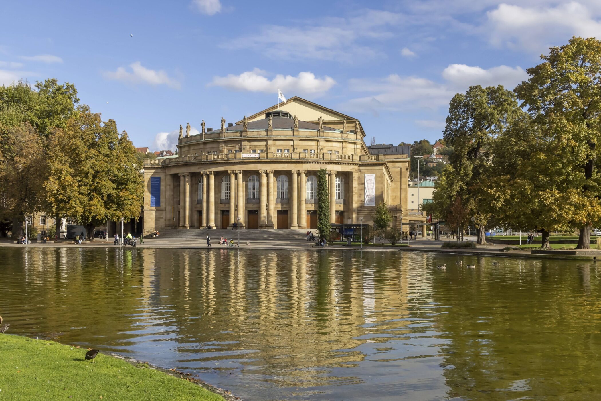Rundes Gebäude mit Säulen, davor ein Teich, umgeben von Bäumen und Menschen.