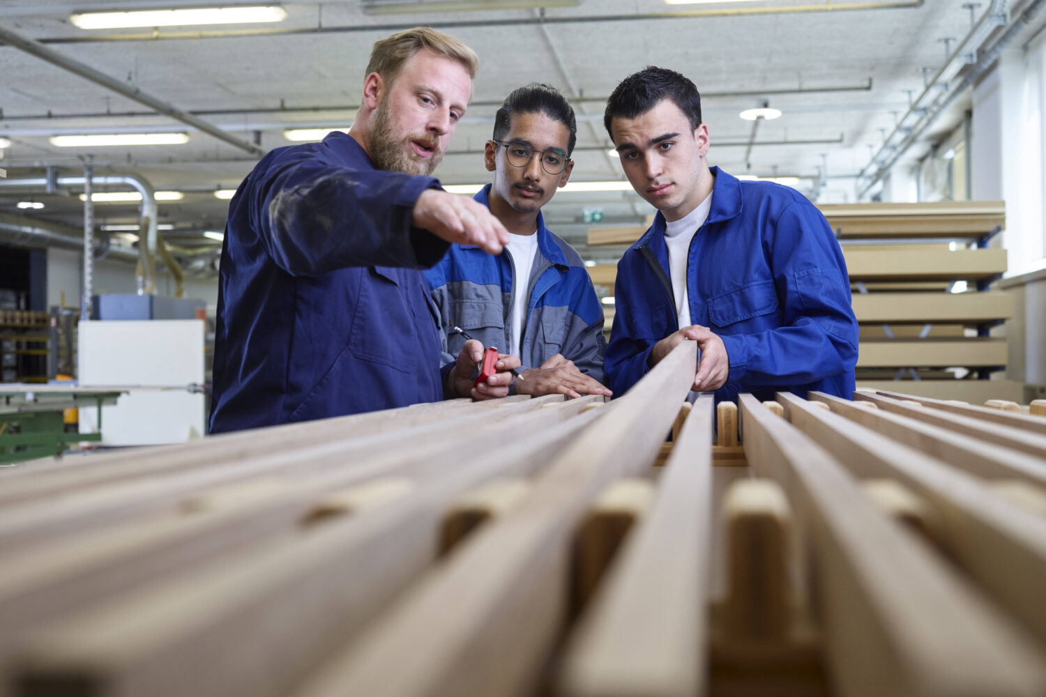Drei Männer in Arbeitskleidung begutachten Holzleisten in einer Werkstatt.