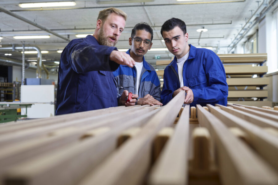 Drei Männer in Arbeitskleidung begutachten Holzleisten in einer Werkstatt.