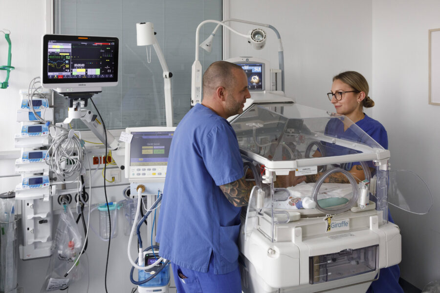 Zwei Personen in blauer Kleidung bei einem Inkubator im Krankenhaus.