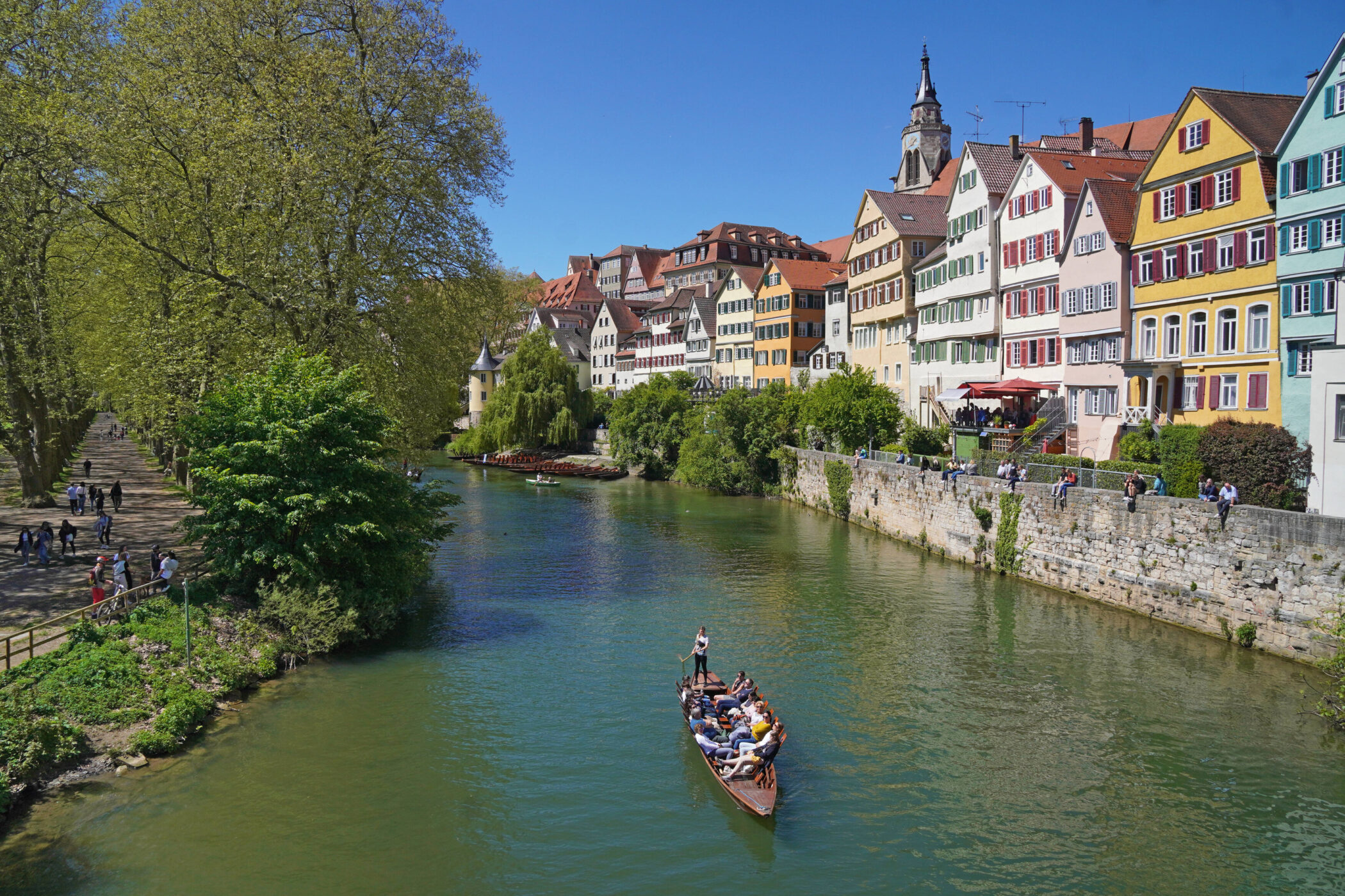 Fluss mit Boot, bunte Häuserreihe rechts, Spaziergänger links unter Bäumen.