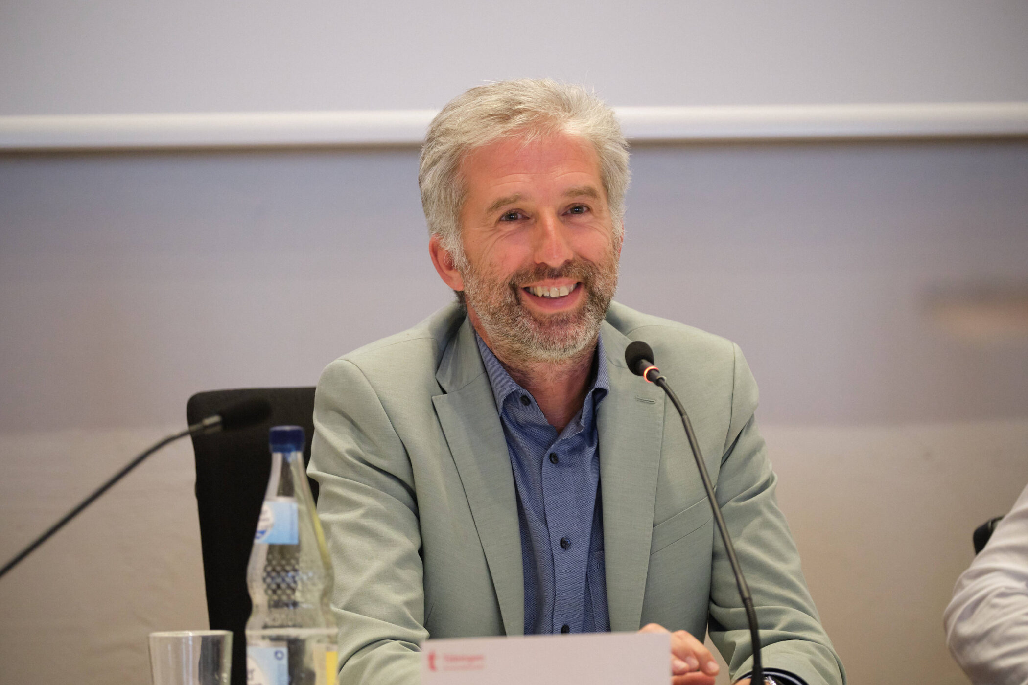 Ein Mann mit grauem Haar spricht in ein Mikrofon, vor ihm eine Wasserflasche.