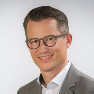 Ein Mann mit Brille und Anzug lächelt vor neutralem Hintergrund.