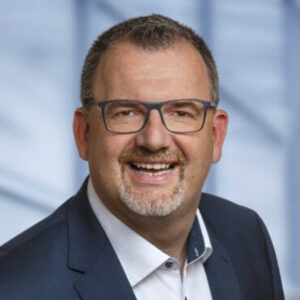 Ein lächelnder Mann mit Brille in Anzug vor unscharfem Hintergrund.