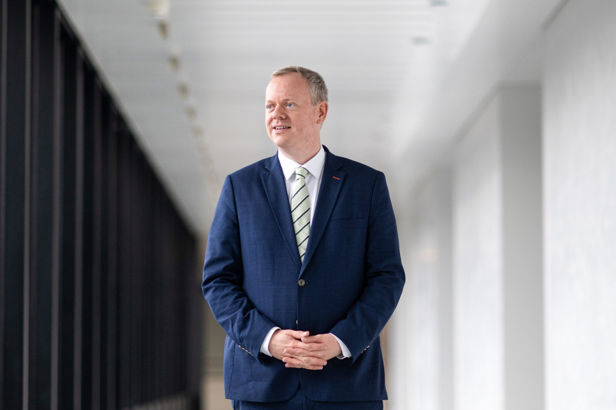 Mann in Anzug steht in einem hellen Flur und schaut zur Seite.