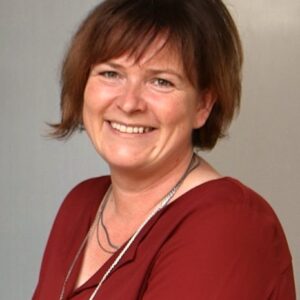 Eine lächelnde Frau in roter Bluse vor grauem Hintergrund.