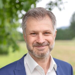 Ein Mann im Anzug lächelt im Freien, Bäume im Hintergrund.