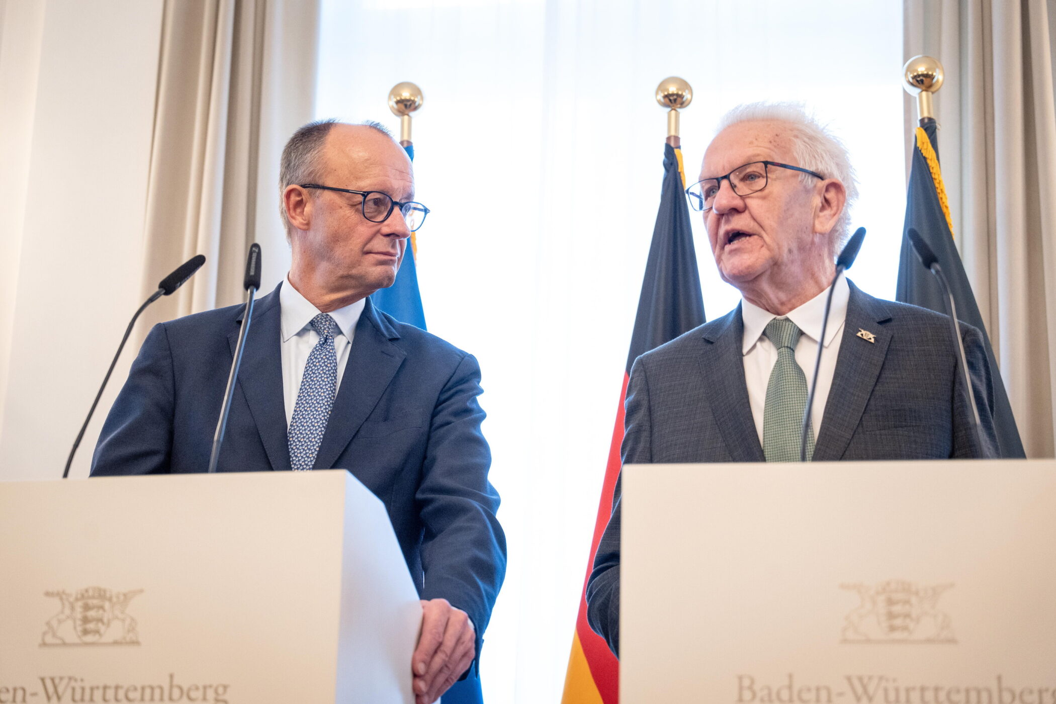 Zwei Männer in Anzügen stehen hinter Rednerpulten mit Mikrofonen, Flaggen im Hintergrund.