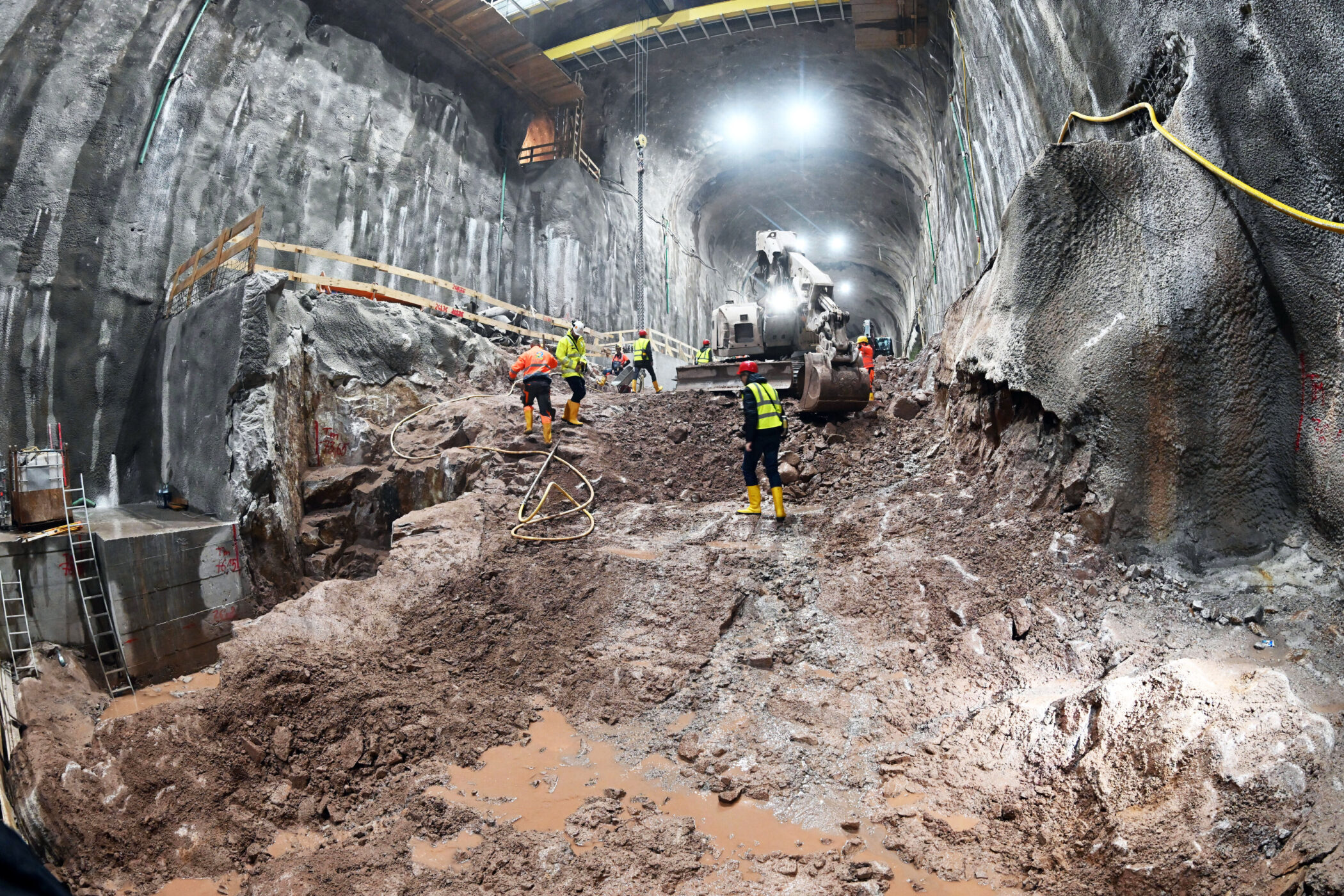 Mehrere Bauarbeiter in einem großen Tunnel mit Bagger und schlammigem Boden.