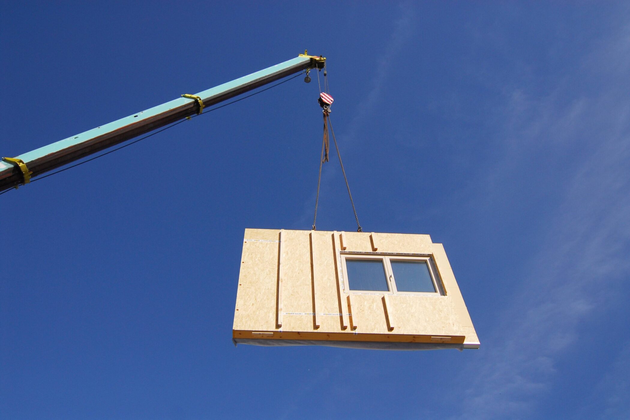 Kran hebt Holzwand mit Fenster vor blauem Himmel.