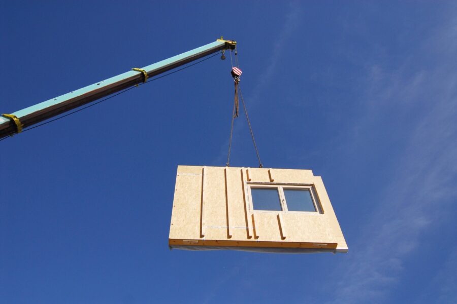Kran hebt Holzwand mit Fenster vor blauem Himmel.