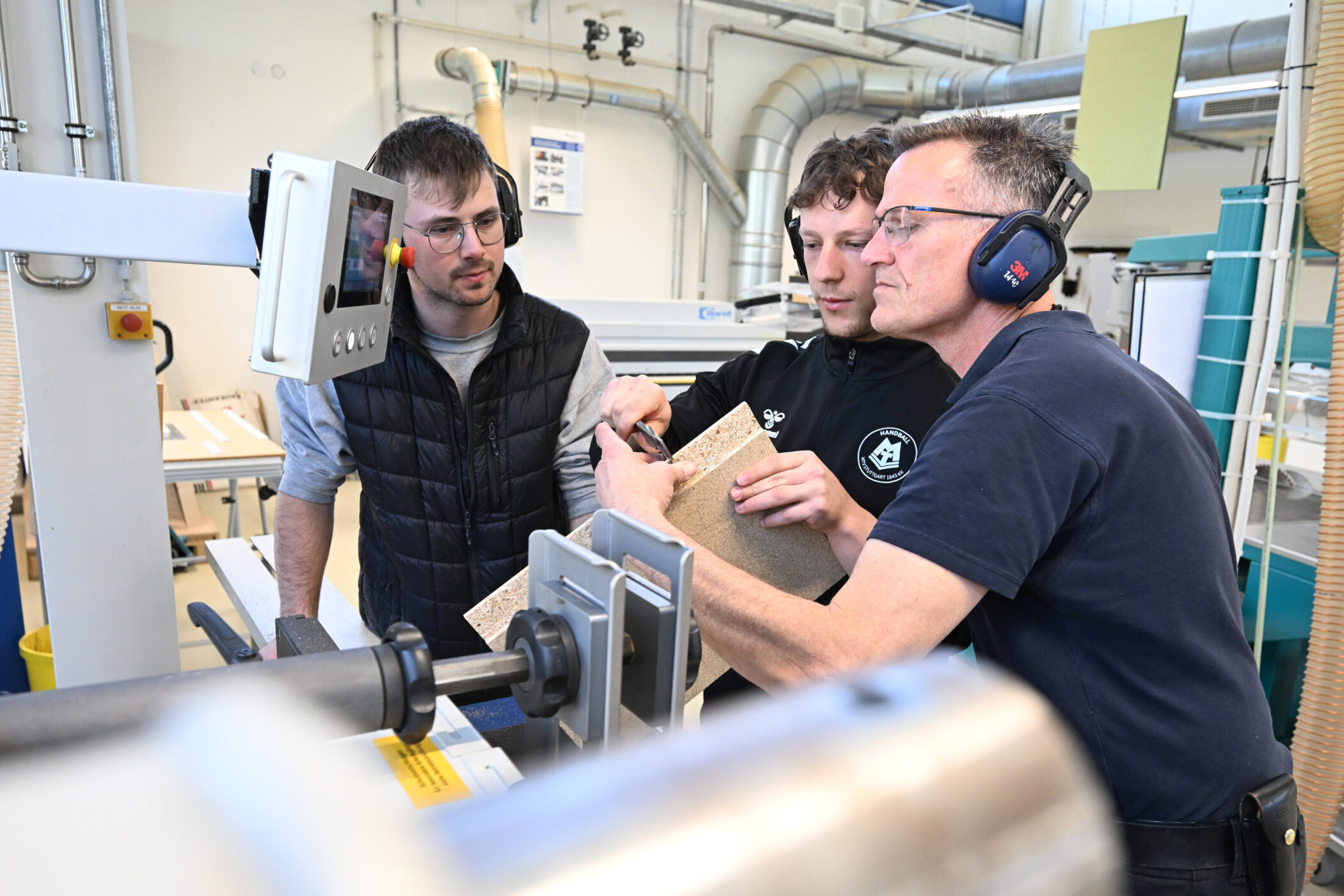 Drei Männer arbeiten in einer Werkstatt an einer Maschine mit einem Holzbrett.