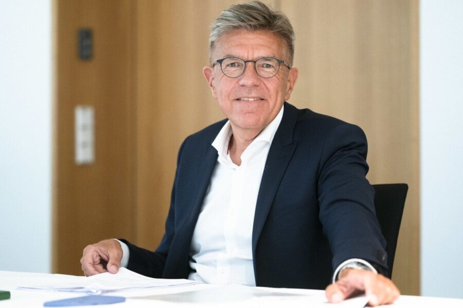 Älterer Mann mit Brille und Anzug sitzt an einem Tisch, lächelt in die Kamera.