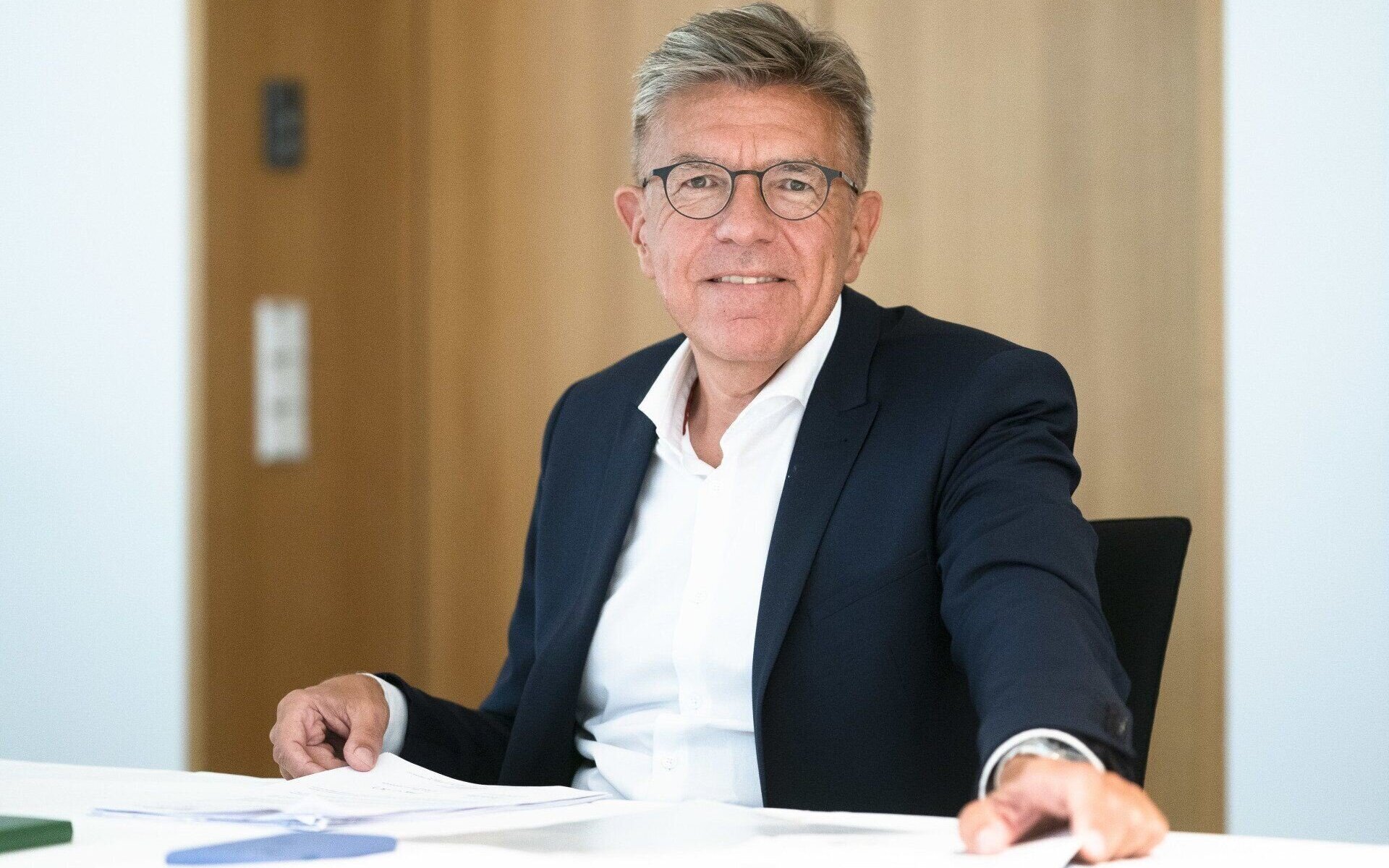 Älterer Mann mit Brille und Anzug sitzt an einem Tisch, lächelt in die Kamera.